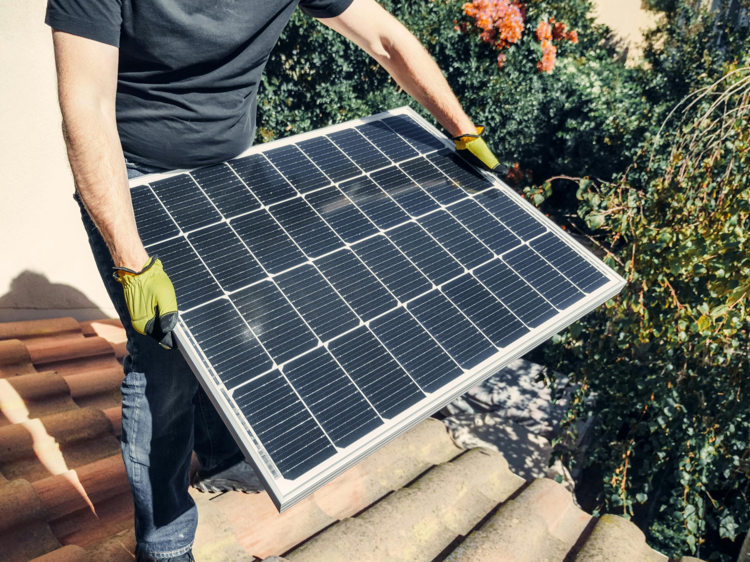 A worker in gloves installs a solar panel on a tiled rooftop, promoting renewable energy.